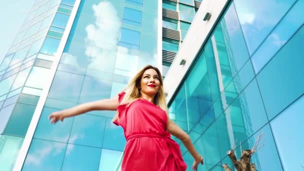 超高層ビルを通り過ぎる赤いドレスを着た美しいブロンドの女性の低角度ショット — ストック動画