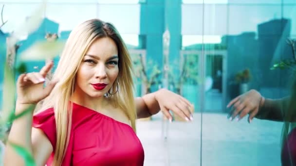 Tanzende blonde Frau in rotem Kleid in der Nähe moderner Gebäude in der Innenstadt — Stockvideo