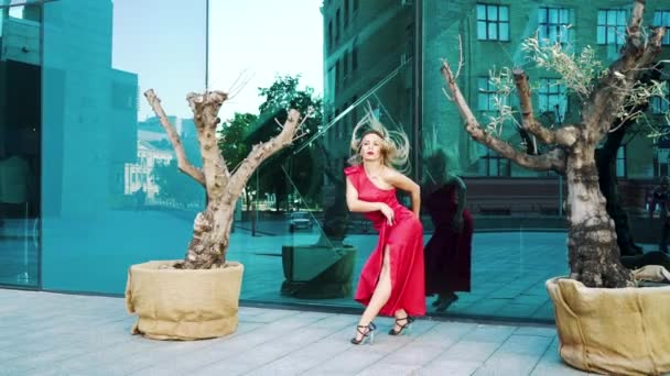 Seductora mujer rubia en vestido rojo bailando cerca de superficies espejo de rascacielos — Vídeo de stock