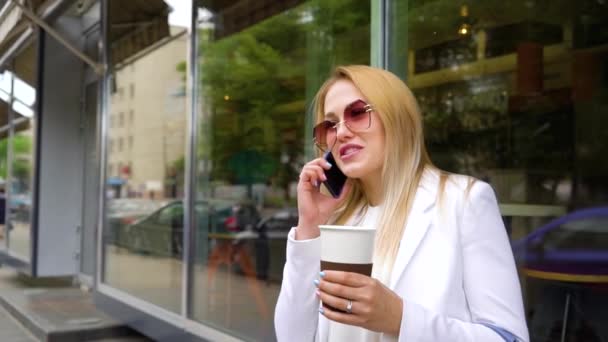 Vacker blond kvinna talar på mobiltelefon som innehar kaffekopp på gatan — Stockvideo