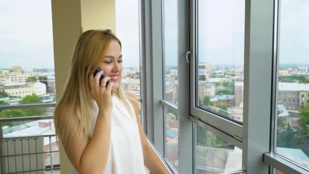 Mujer joven habla en el teléfono móvil en la habitación espaciosa con paisaje urbano en el fondo — Vídeo de stock