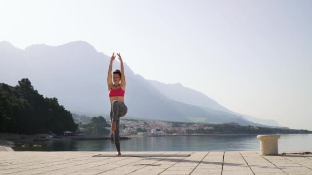 Wanita dewasa fit melakukan yoga di dermaga laut kosong dengan pegunungan di latar belakang — Stok Video
