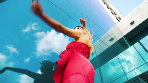 Низкий угол сексуальной блондинки танцующей против стеклянного здания, отражающего небо — стоковое видео