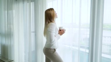 genç sarışın kadın bir fincan ile büyük perdeli pencere yakınında rahat kalan