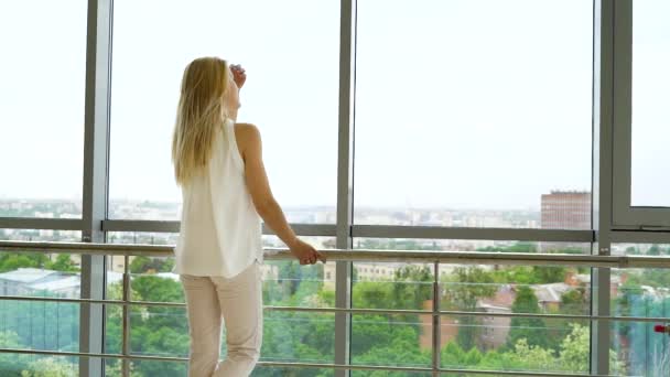 Mujer joven y elegante con el pelo largo y rubio mirando por la ventana grande de la ciudad — Vídeos de Stock