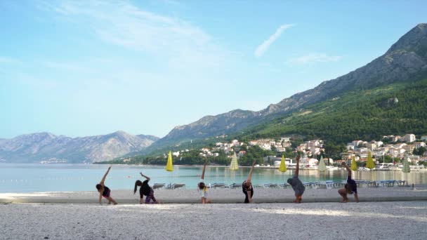 Группа молодых людей, стоящих в асанах для йоги на пустом морском пляже утром — стоковое видео
