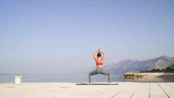 Visão traseira da mulher fazendo ioga à beira-mar com montanhas e céu no fundo — Vídeo de Stock