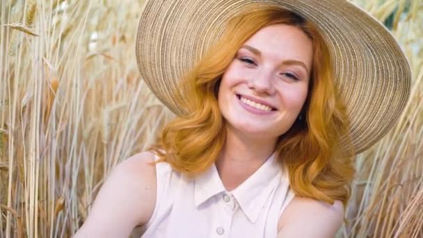 Портрет молодої рудої волохатої жінки в солом'яному капелюсі, що посміхається на камеру в пшеничному полі — стокове відео