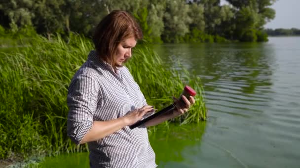 Женщина-эколог с планшетом изучает образец зеленых водорослей на берегу реки — стоковое видео