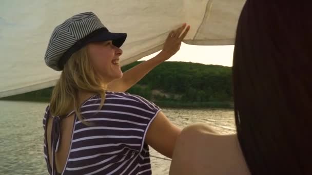 Дві веселі молоді жінки відпочивають на вітрильному човні, що плаває на річці на заході сонця — стокове відео