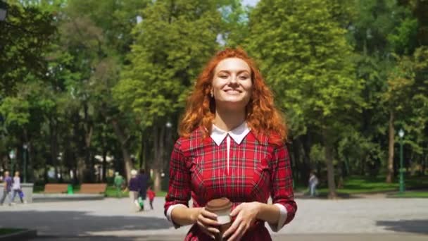 Chica pelirroja positiva caminando en el parque y tomando café — Vídeos de Stock