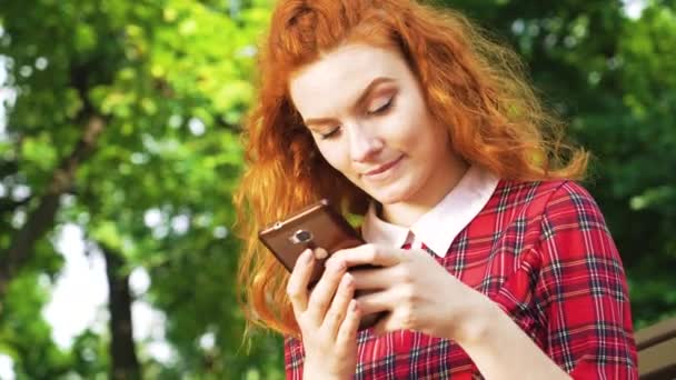 Felice ragazza carina con i capelli rossi digitando messaggio sullo smartphone seduto nel parco — Video Stock