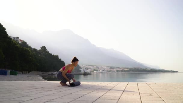 Donna atletica in abbigliamento sportivo che srotola tappetino yoga sul molo del mare — Video Stock