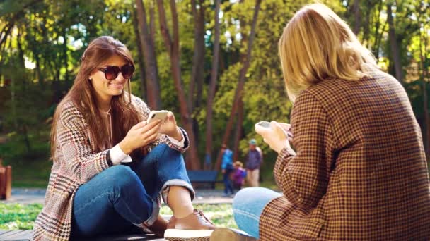 Chicas sin emociones adictas con teléfonos inteligentes en el soleado parque — Vídeo de stock