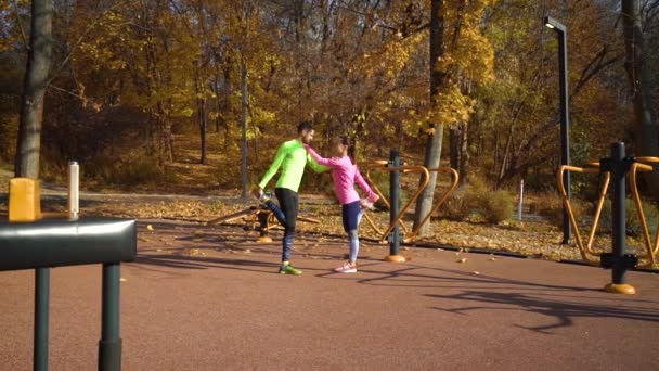 Ajuste pareja sosteniéndose y estirando las piernas en el campo de deportes en otoño — Vídeos de Stock