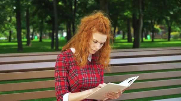 Unhappy red haired girl reading thriller in park — ストック動画
