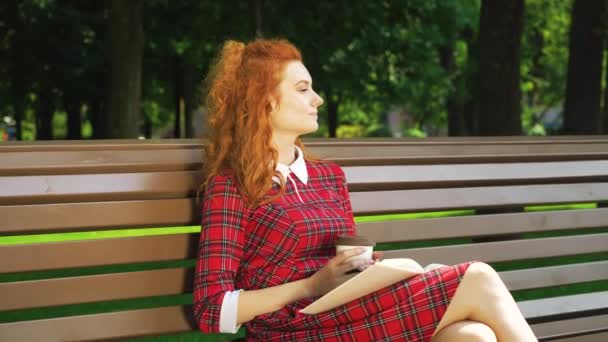 Όμορφη κοκκινομάλλα κορίτσι χαλάρωση στο πάρκο με το βιβλίο και τον καφέ — Αρχείο Βίντεο