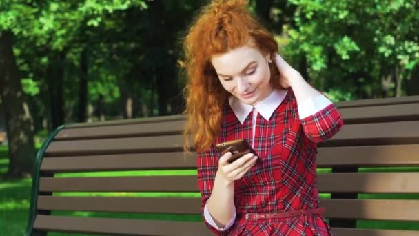 Imádnivaló vörös hajú lány gépelés üzenet okostelefon a zöld parkban — Stock videók