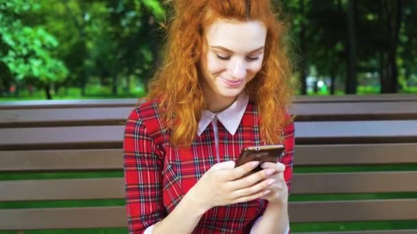 Positive redhead girl texting message on phone in green park — Stock Video