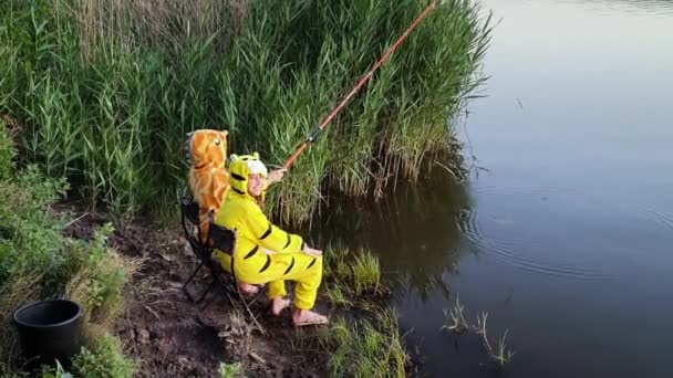 Gadis lucu dengan piyama lucu duduk dengan batang ikan di tepi sungai — Stok Video
