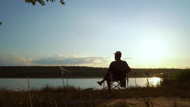 Gün batımında nehir kıyısında sandalyede oturan balıkçı silueti. — Stok video