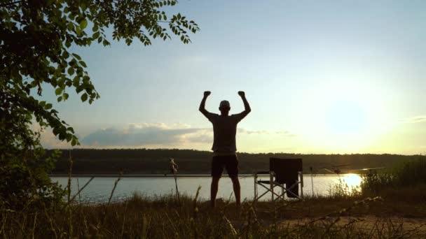 Silhouette of fisherman expressing joyful feelings on river bank at sunset — Stock Video