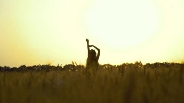 Gün batımında buğday tarlasında kolları havada duran kız silueti.