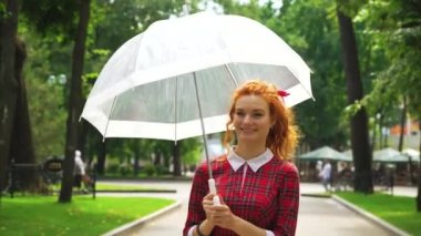 Kızıl saçlı kız güneşli bir günde elinde şemsiyeyle parkta yürüyor.