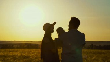 Anne ve babanın siluetleri gün batımında buğday tarlasında bebeği tutuyor.