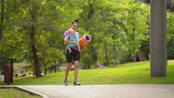 Мать Йоги носит ребенка в стропе перед тренировкой в зеленом парке — стоковое видео