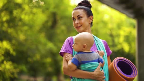 Sportliche Mutter mit Baby im Tragetuch zu Fuß zum Yoga-Kurs im grünen Park — Stockvideo