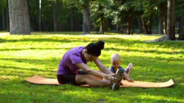 Kadın bacak esnetiyor ve bebek parkta mindere oturuyor.