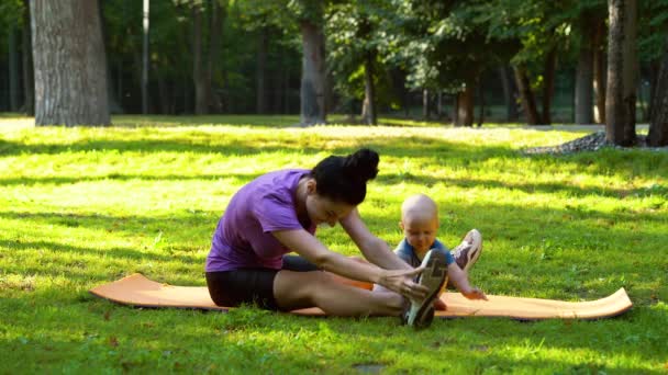 Жінка розтягує ноги і дитина сидить поруч з нею на килимку в парку — стокове відео