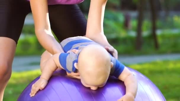 Madre haciendo ejercicios de gimnasia con el bebé en la pelota de fitness en el parque — Vídeo de stock