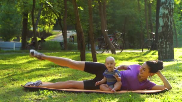 Młoda kobieta ćwiczy i słodkie dziecko siedzi obok niej w parku — Wideo stockowe