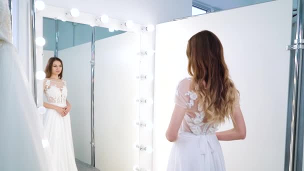 Молодая женщина смотрит в зеркало в свадебном салоне — стоковое видео