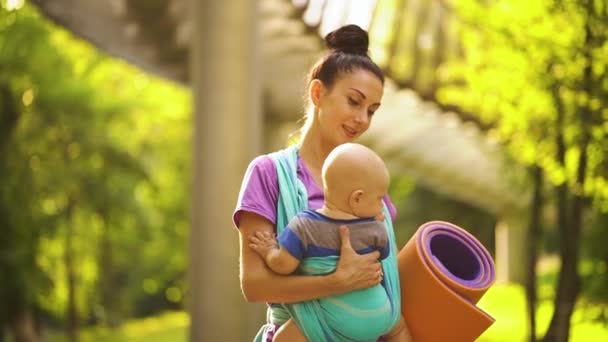 Femme sportive portant bébé en fronde et marchant jusqu'au cours de yoga dans le parc — Video