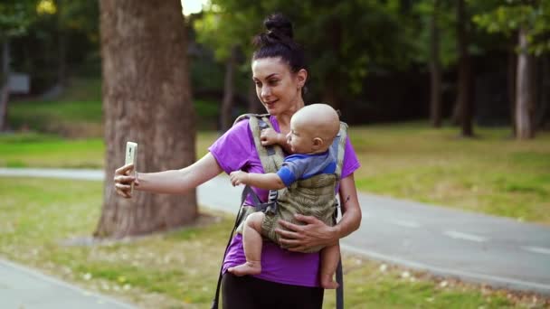 Młoda kobieta robi selfie podczas noszenia dziecka w temblaku — Wideo stockowe