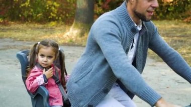 Baba kızıyla bisiklete biniyor, çocuk koltuğunda oturuyor.