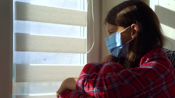 Девушка в маске сидит дома и смотрит в окно — стоковое видео