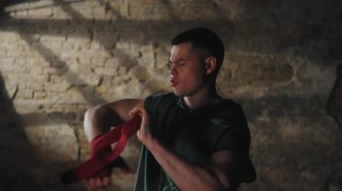 Yeşil kapüşonlu kaslı adam kırmızı boks elini bileğine dolayıp loş spor salonunda tuğladan duvarlar ve dramatik bir şekilde aydınlatıyor.