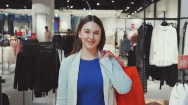 Giyim mağazasında parlak bir alışveriş çantasıyla dolaşan, kameraya gülümseyen ve modern moda perakende deneyiminin tadını çıkaran mutlu genç bir kadın.
