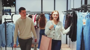 Genç çift mağazada alışveriş yapmaktan hoşlanıyor, kadın çanta tutuyor ve elbiselerini karıştırıyor, erkek ise yanında alışverişlerle yürüyor.