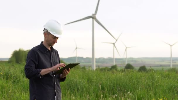 Arbetare står nära vita vindkraftverk, att skriva på en surfplatta. Väderkvarnar, grön energikoncept. — Stockvideo