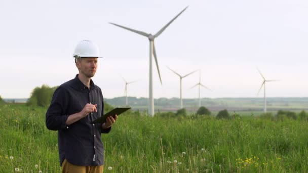 El trabajador se para cerca de los aerogeneradores blancos, escribiendo en una tableta. Molinos de viento, concepto de energía verde . — Vídeos de Stock