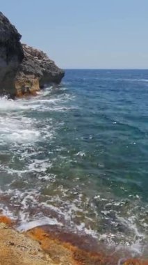 Dalgalar yavaşça kayalık kıyılara, berrak mavi suya ve köpüklü sörflere çarpar. Dikey video.