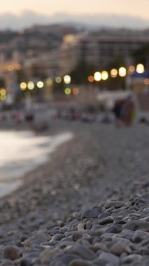 Sahildeki pürüzsüz taşların yakın çekimi, şehrin ışıkları uzaktan yanıp sönüyor. Dikey video.