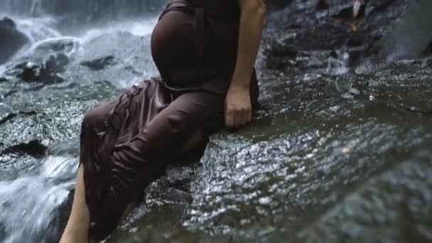 Тяжело беременная женщина, сидящая на скале возле водопада, наслаждается прекрасным видом на природу. Медленное движение . — стоковое видео