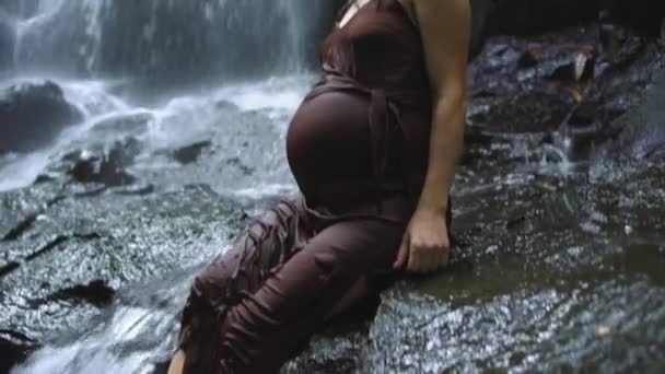 Ευτυχισμένη έγκυος γυναίκα κάθεται σε ένα βράχο κοντά στον καταρράκτη και κοιτάζοντας την κάμερα. Αργή κίνηση. — Αρχείο Βίντεο
