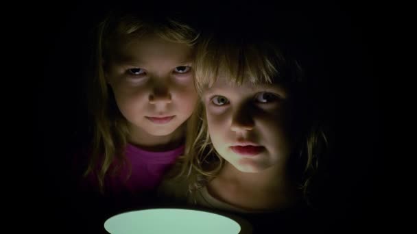 Nahaufnahme zwei kleine Mädchen machen beängstigende Gesichter, die von unten in einem dunklen Raum eine Lampe auf sich selbst scheinen lassen — Stockvideo